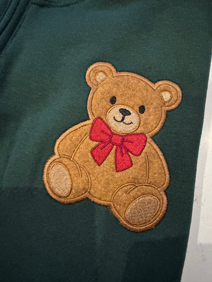 Close-up of a brown teddy bear applique with plush textured fabric, embroidered outlines and a bright pink bow on a forest green sweatshirt.