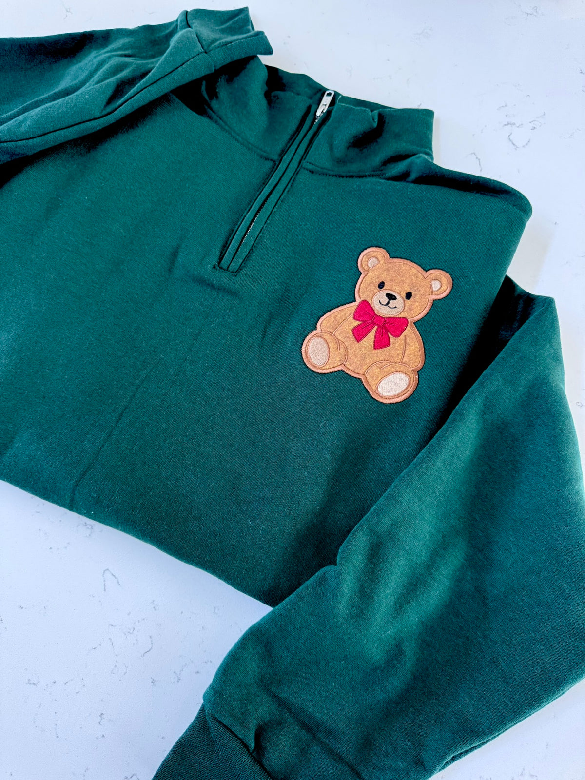 A dark green quarter-zip hoodie featuring an embroidered brown teddy bear with a red bow on the left chest, laid flat on a light textured surface.