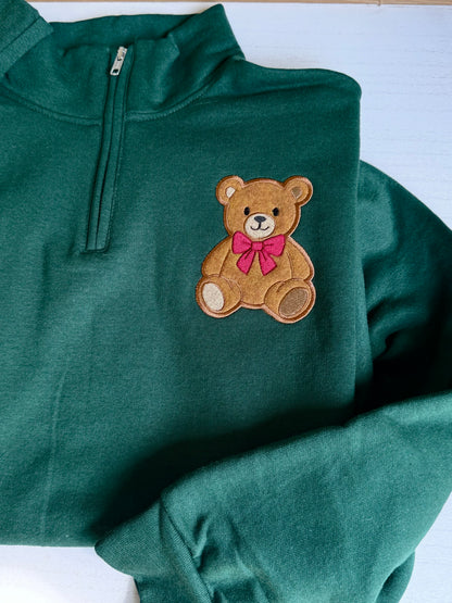 Close-up of a forest green quarter zip pullover showing the embroidered teddy bear applique, red bow and soft sweatshirt fabric.