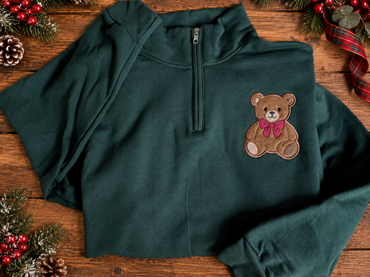 Forest green quarter zip sweatshirt with embroidered teddy bear and red bow displayed on rustic wooden planks with Christmas greenery and red berries.