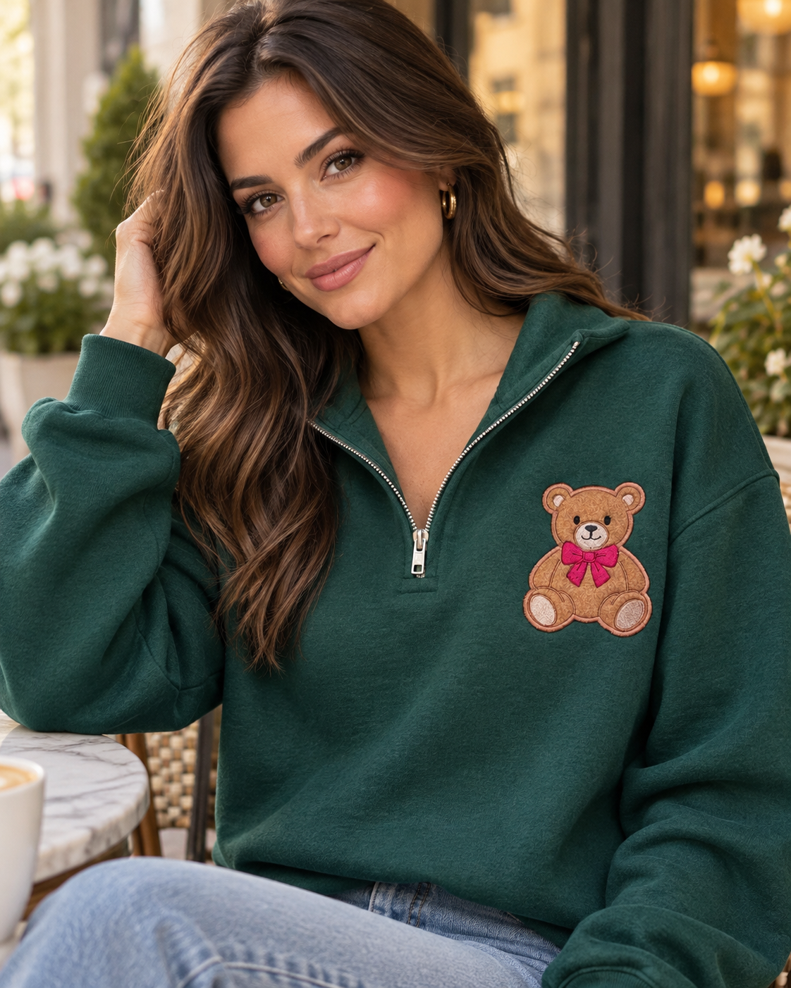 Brunette woman wearing a forest green quarter zip teddy bear sweatshirt with jeans at an outdoor cafe, styled for a cozy chic look.
