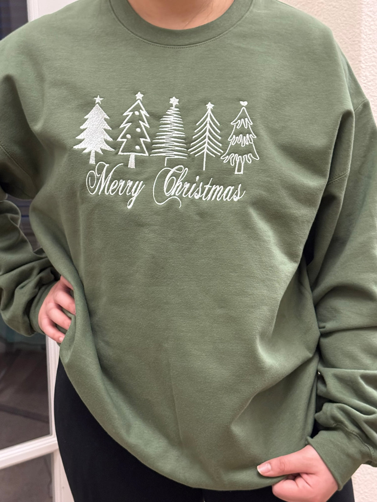 A person wearing an heather green crewneck sweatshirt with a white thread embroidered of five Christmas trees and the words 'Merry Christmas' embroidered on the front.