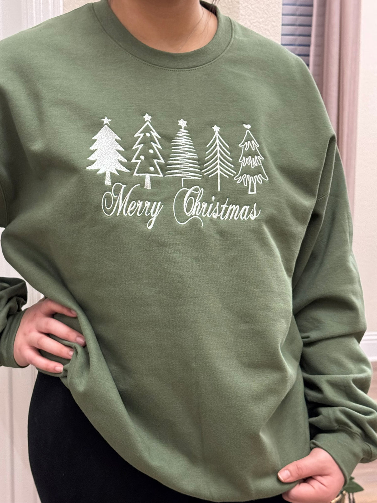 woman wear the heather green sweatshirt with  white thread Merry Christmas embroidery with four festive Christmas trees,