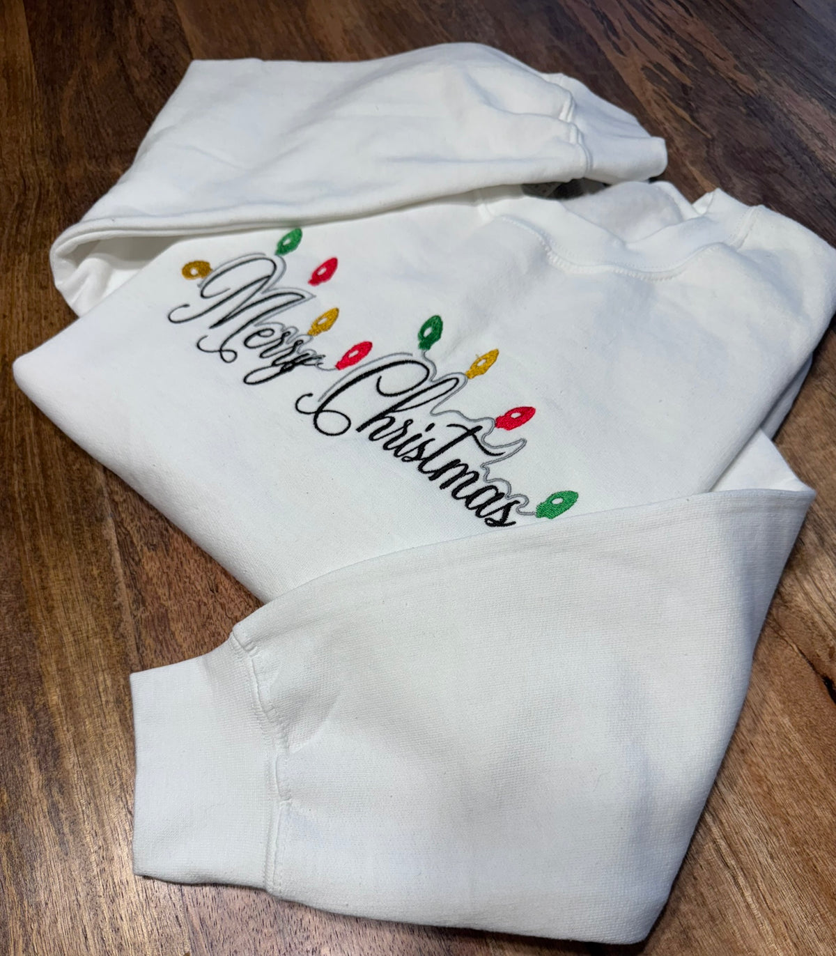 A white sweatshirt with 'Merry Christmas' written in black script, decorated with colorful Christmas lights, laid flat on a dark wood background.