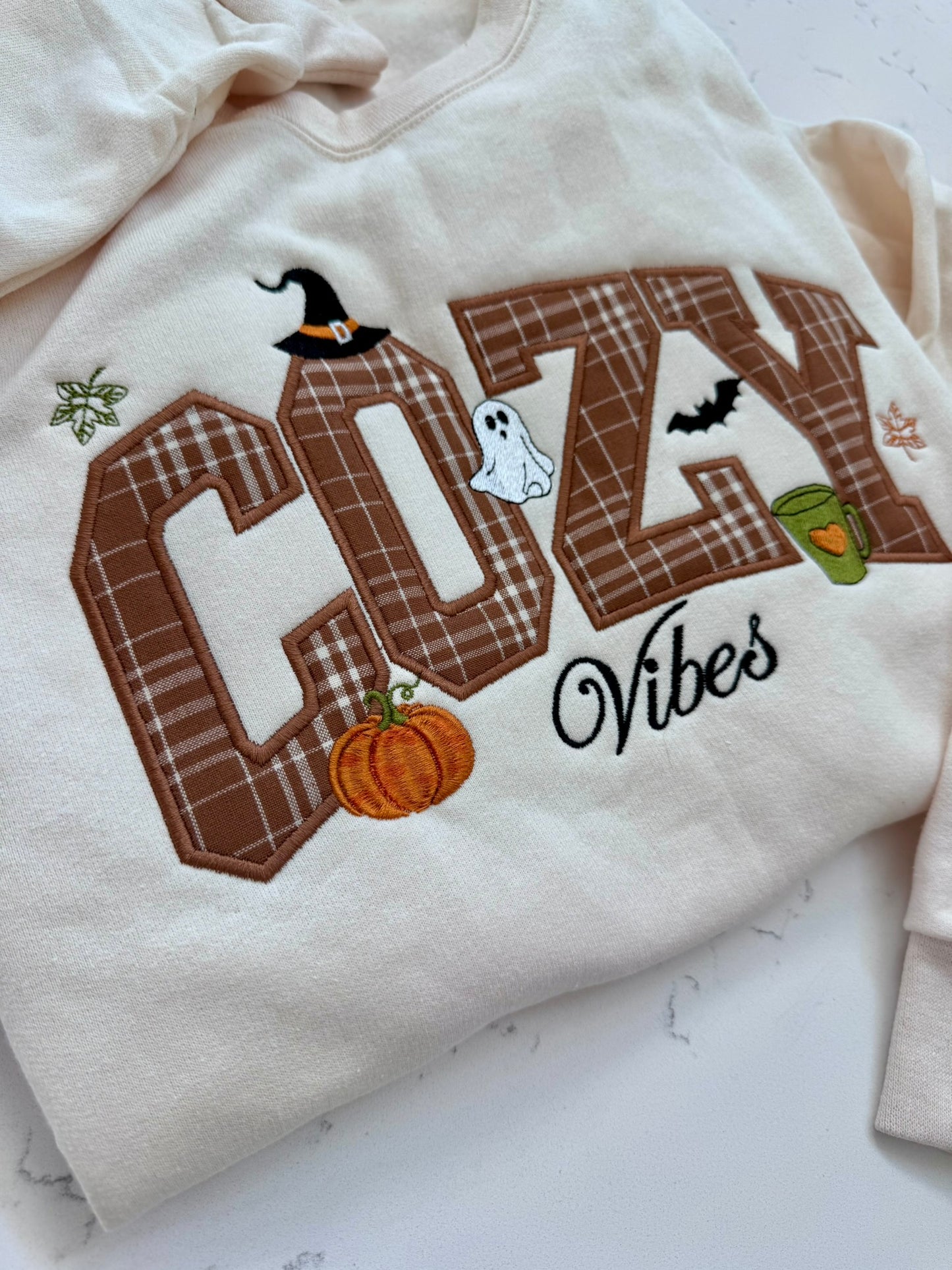 A cream-colored sweatshirt featuring a large embroidered 'COZY Vibes' design with autumn Halloween motifs including a witch hat, ghost, pumpkin, bat, and green cup, laid flat on a light marble surface.