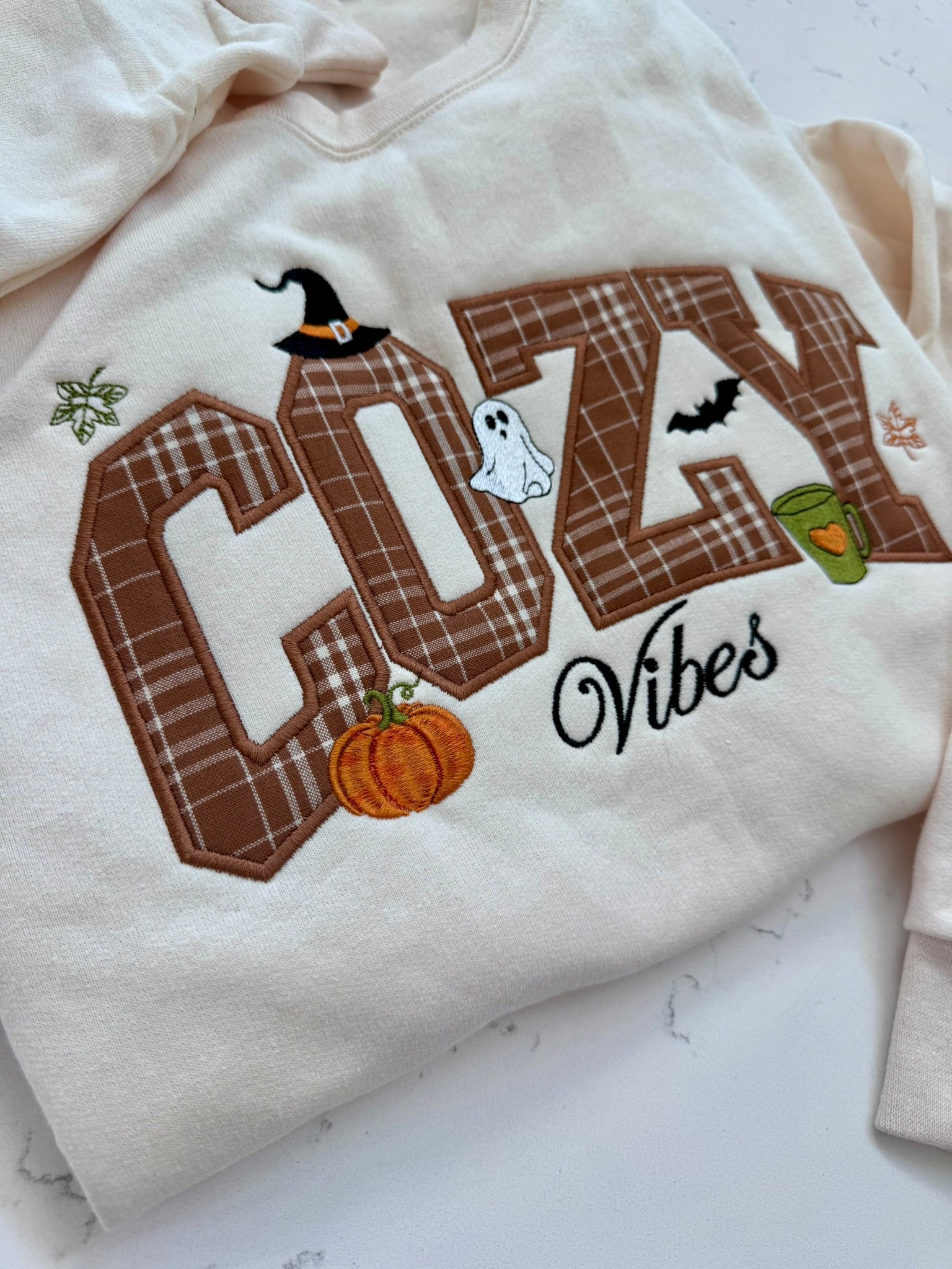 A cream-colored sweatshirt featuring a large embroidered 'COZY Vibes' design with autumn Halloween motifs including a witch hat, ghost, pumpkin, bat, and green cup, laid flat on a light marble surface.