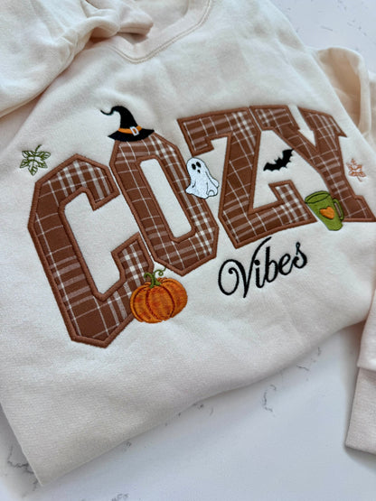 A cream-colored sweatshirt featuring a large embroidered 'COZY Vibes' design with autumn Halloween motifs including a witch hat, ghost, pumpkin, bat, and green cup, laid flat on a light marble surface.