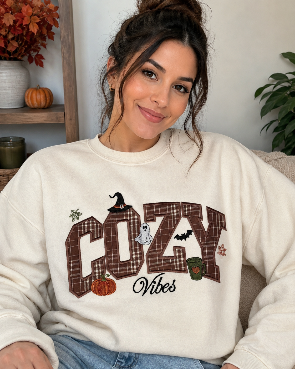 A woman wearing a cream-colored crewneck sweatshirt with a 'COZY Vibes' autumn graphic, sitting in a cozy indoor setting.