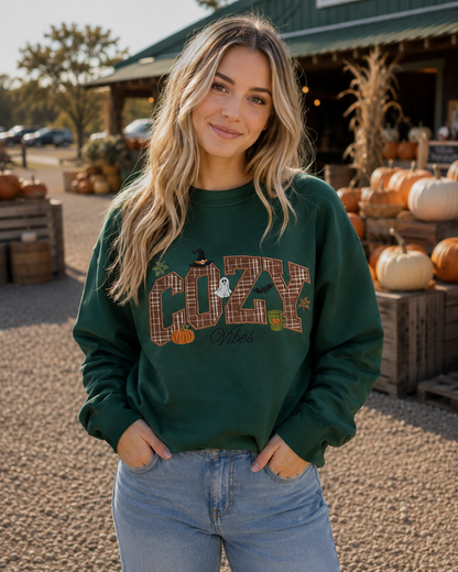 A woman stands outdoors in front of a fall farm stand, wearing a dark green crewneck sweatshirt with a 'COZY Vibes' autumn graphic and light wash blue jeans.