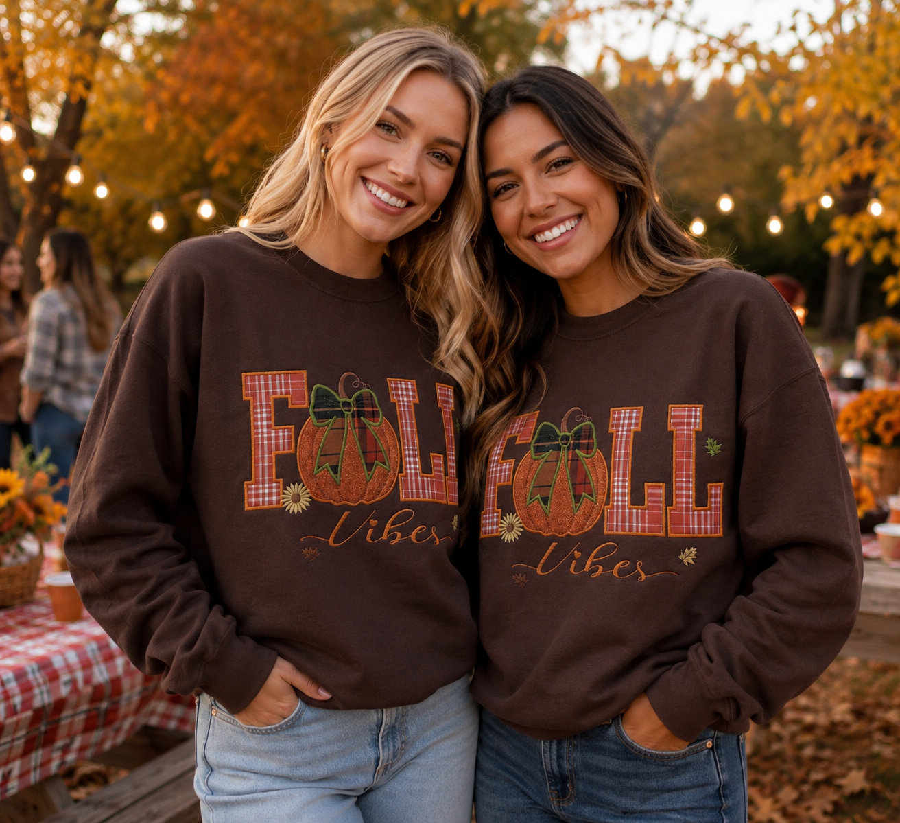 Two women standing outdoors in a fall-themed setting, wearing matching dark brown sweatshirts with a 'FALL Libes' graphic featuring pumpkins and gingham lettering.