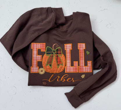A dark brown crewneck sweatshirt featuring a fall-themed graphic that reads 'FALL Vibes' with a plaid pumpkin and bow, laid flat on a light textured background.