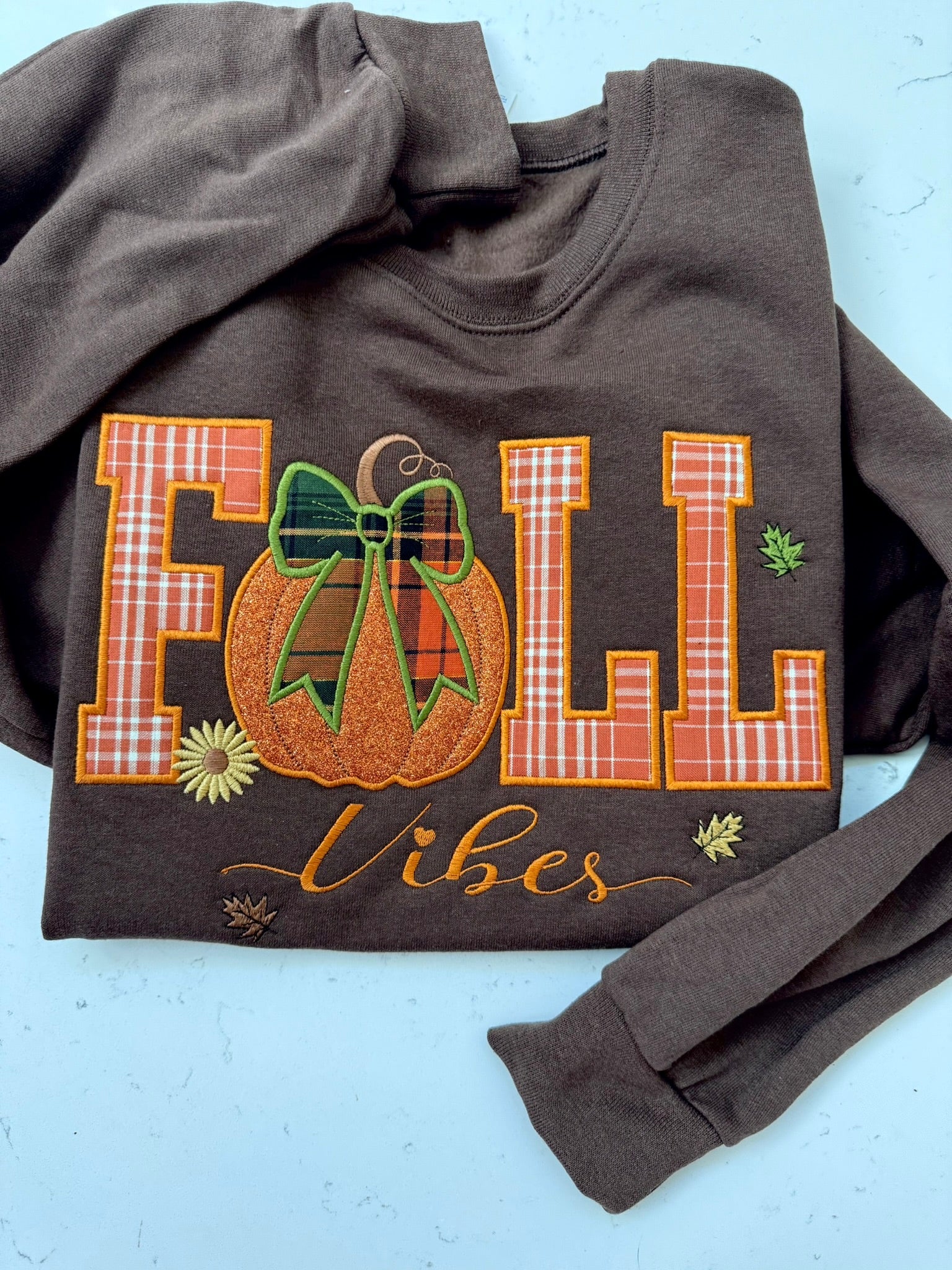 A dark brown crewneck sweatshirt featuring a 'FALL Vibes' embroidered graphic on the front, laid flat on a light blue background.
