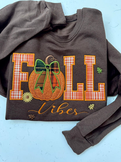 A dark brown crewneck sweatshirt featuring a 'FALL Vibes' embroidered graphic on the front, laid flat on a light blue background.