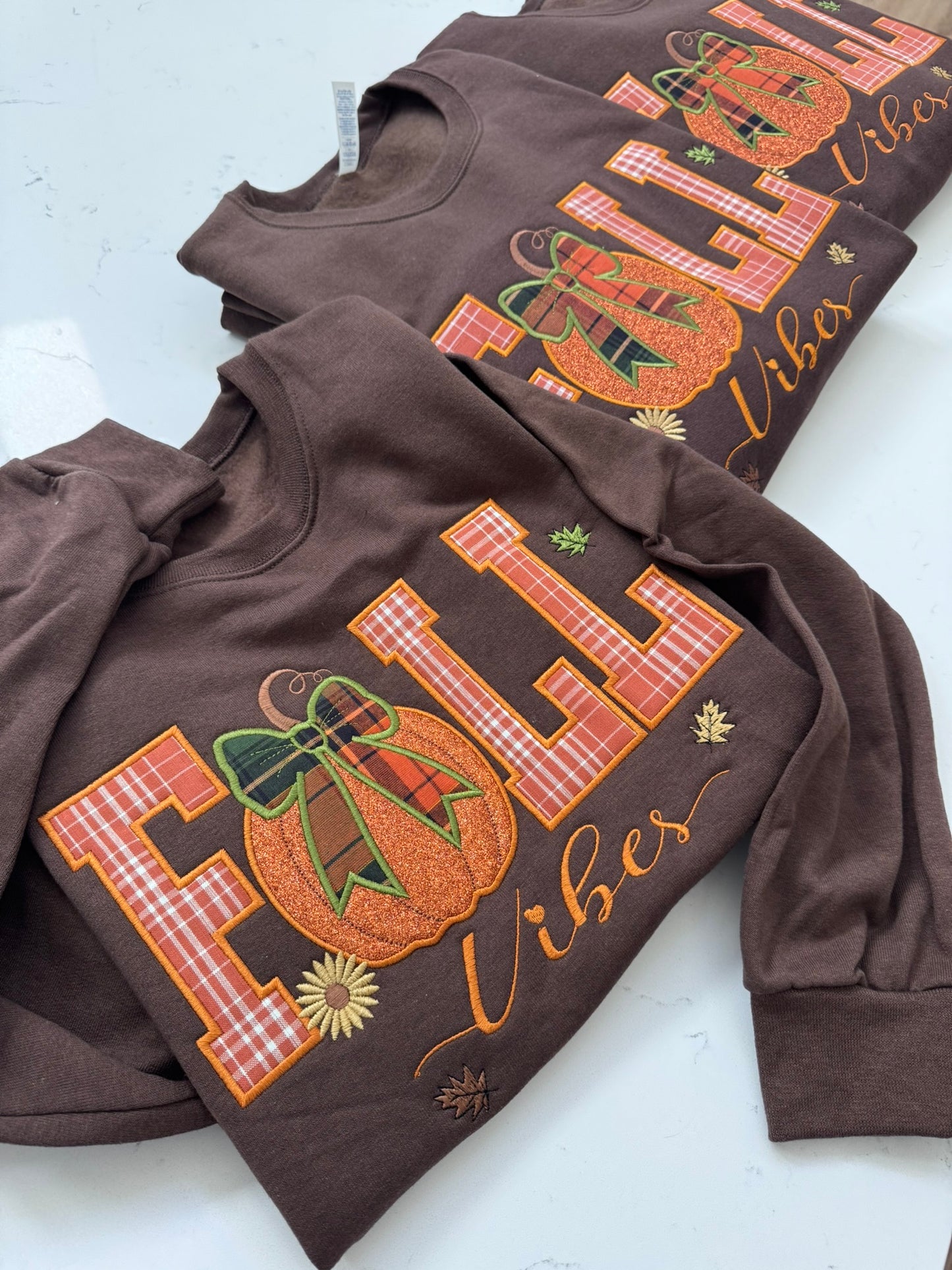 A brown long-sleeve sweatshirt with a large 'FALL Vibes' graphic featuring an orange pumpkin and autumn leaves, laid flat on a white surface with two matching sweatshirts partially visible behind it.