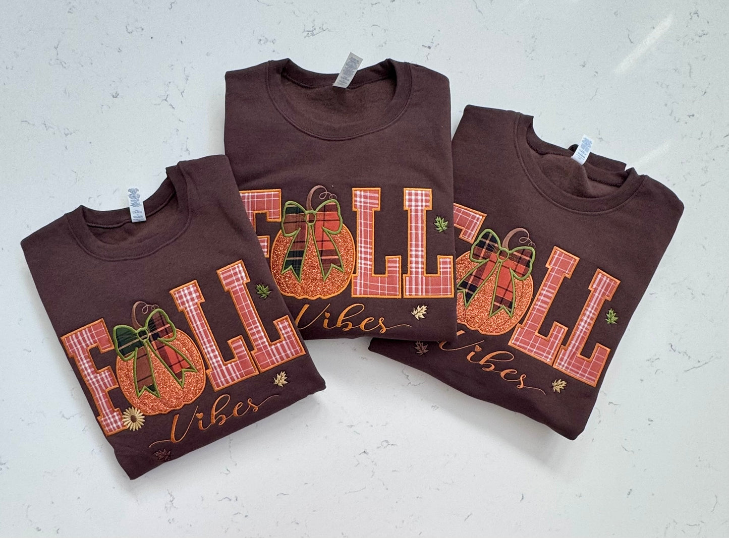 Three folded brown crewneck t-shirts with a fall-themed graphic reading 'FALL vibes' in orange plaid letters, decorated with a pumpkin and bow motif, displayed on a light gray marble surface.