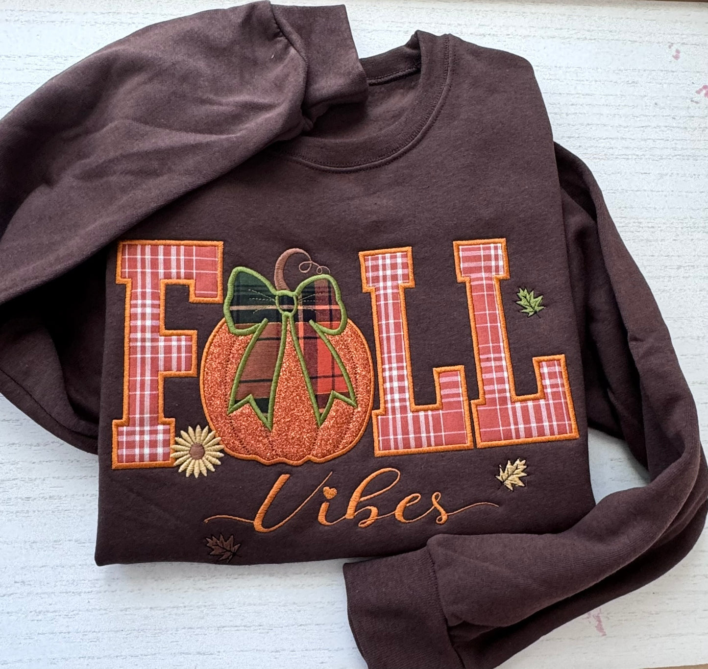 A dark brown crewneck sweatshirt laid flat on a white wood background, featuring a large 'FALL Vibes' embroidery design with a pumpkin and bow motif.