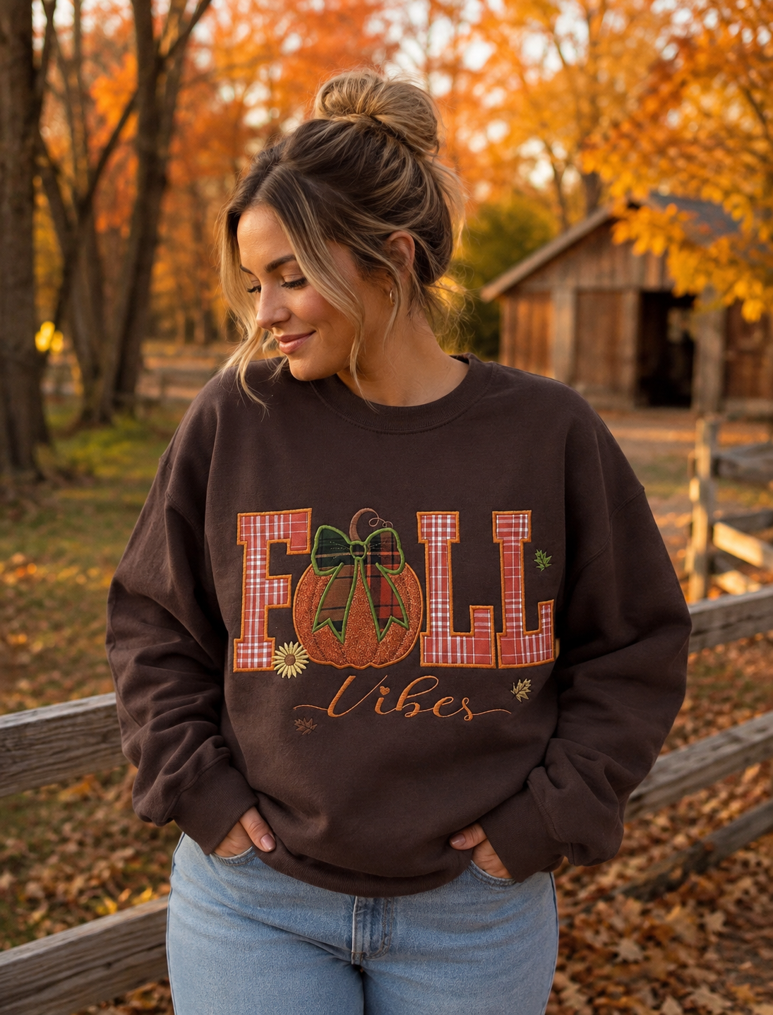 A woman wearing a dark brown crewneck sweatshirt with a 'FALL Vibes' pumpkin graphic, standing outdoors in an autumn park with orange foliage.