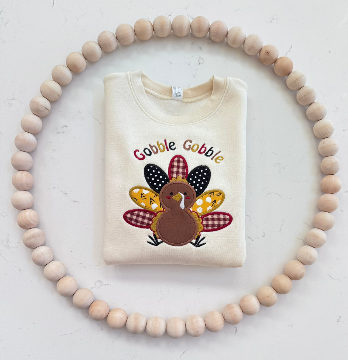 Folded cream toddler Thanksgiving sweatshirt with colorful Gobble Gobble turkey applique embroidery displayed with natural wooden beads.