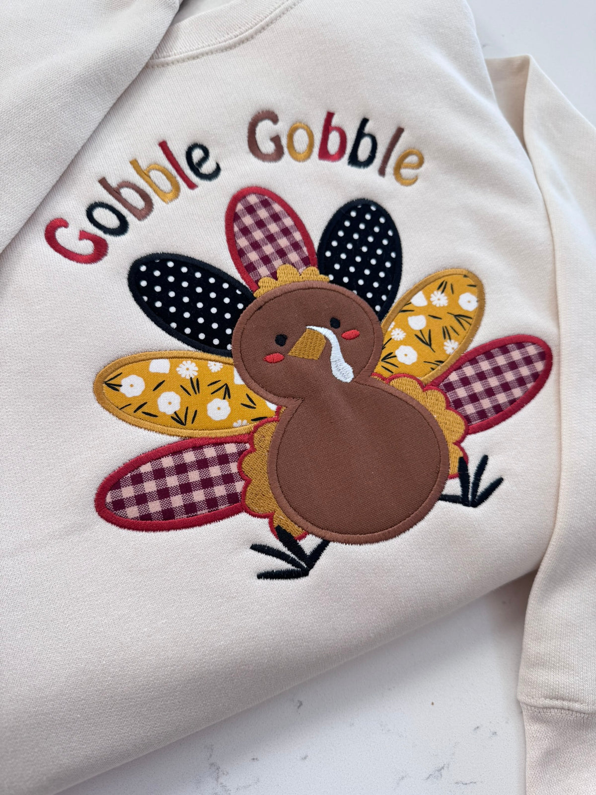Close-up of Gobble Gobble embroidered turkey applique on a cream toddler Thanksgiving sweatshirt featuring colorful patterned fabric feathers.