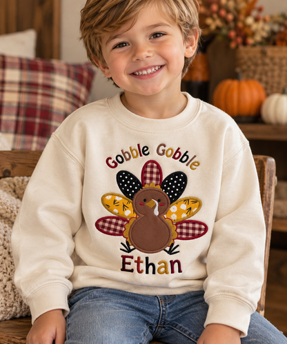 Smiling toddler boy wearing a cream Gobble Gobble Thanksgiving sweatshirt with colorful embroidered turkey applique and personalized name Ethan.