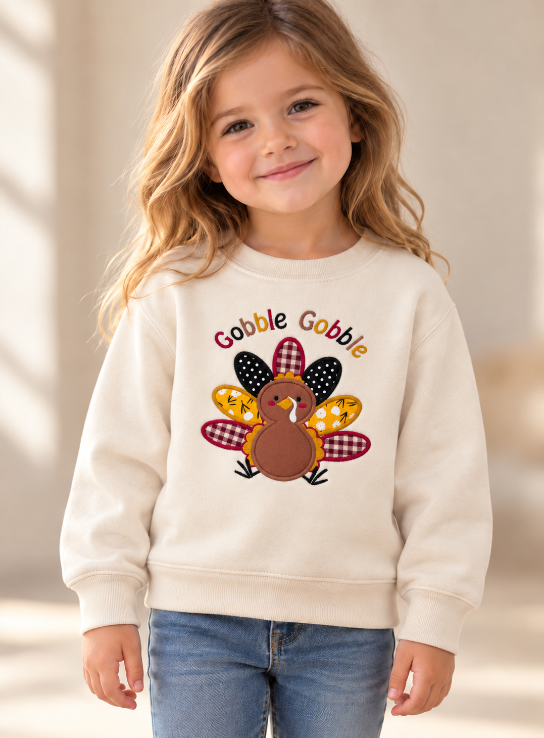 Young girl wearing a cream Gobble Gobble Thanksgiving sweatshirt featuring a colorful embroidered turkey with patterned fabric feathers.