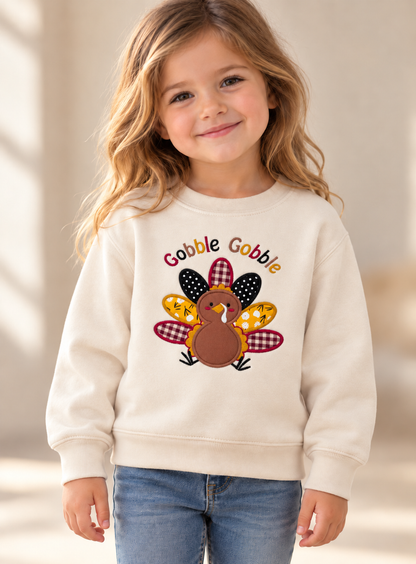 Young girl wearing a cream Gobble Gobble Thanksgiving sweatshirt featuring a colorful embroidered turkey with patterned fabric feathers.