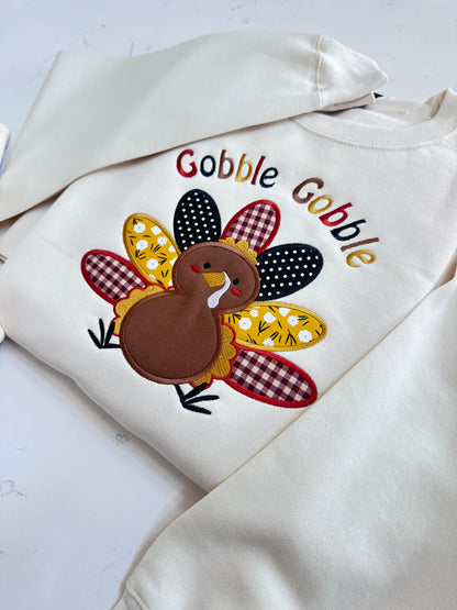 Close-up of cream Gobble Gobble sweatshirt showing colorful turkey applique, embroidered lettering and textured fabric details.