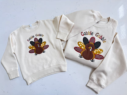 Matching Thanksgiving turkey applique sweatshirts for mom and toddler with colorful embroidery on soft cream crewnecks.