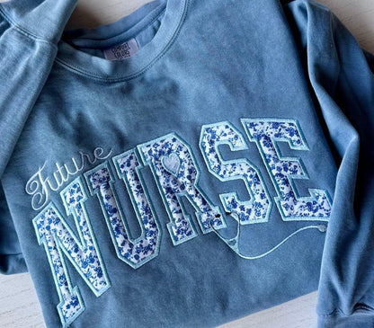 Blue sweatshirt with embroidered 'Future Nurse' text in floral letters on a wooden surface