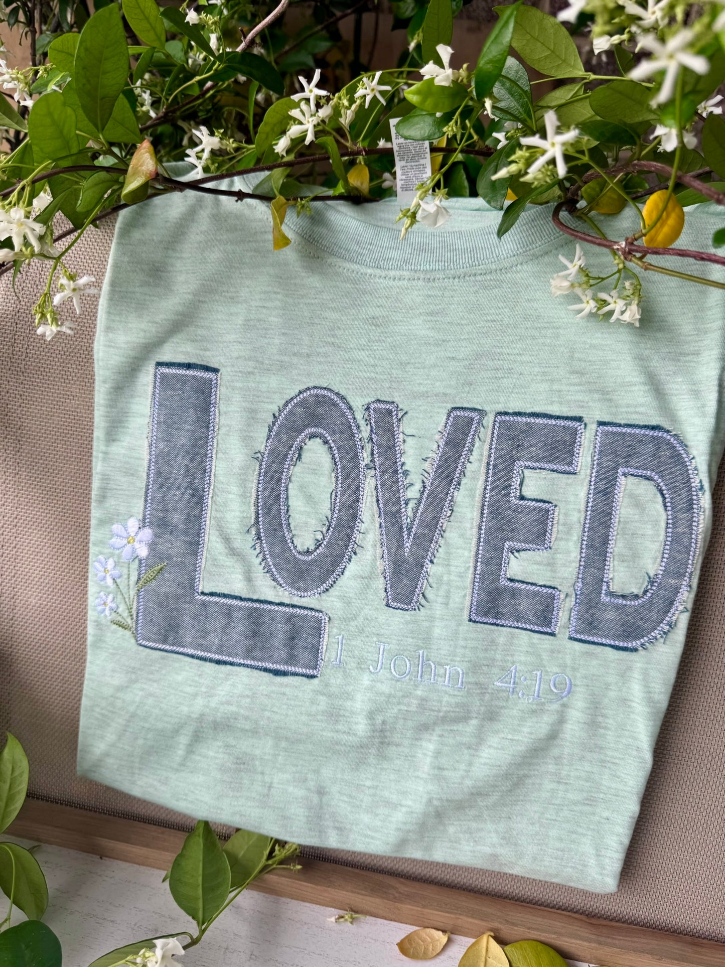 Green t-shirt with 'LOVED' text on a wooden surface with greenery.