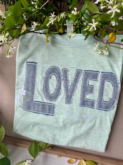Green t-shirt with 'LOVED' text on a wooden surface with greenery.