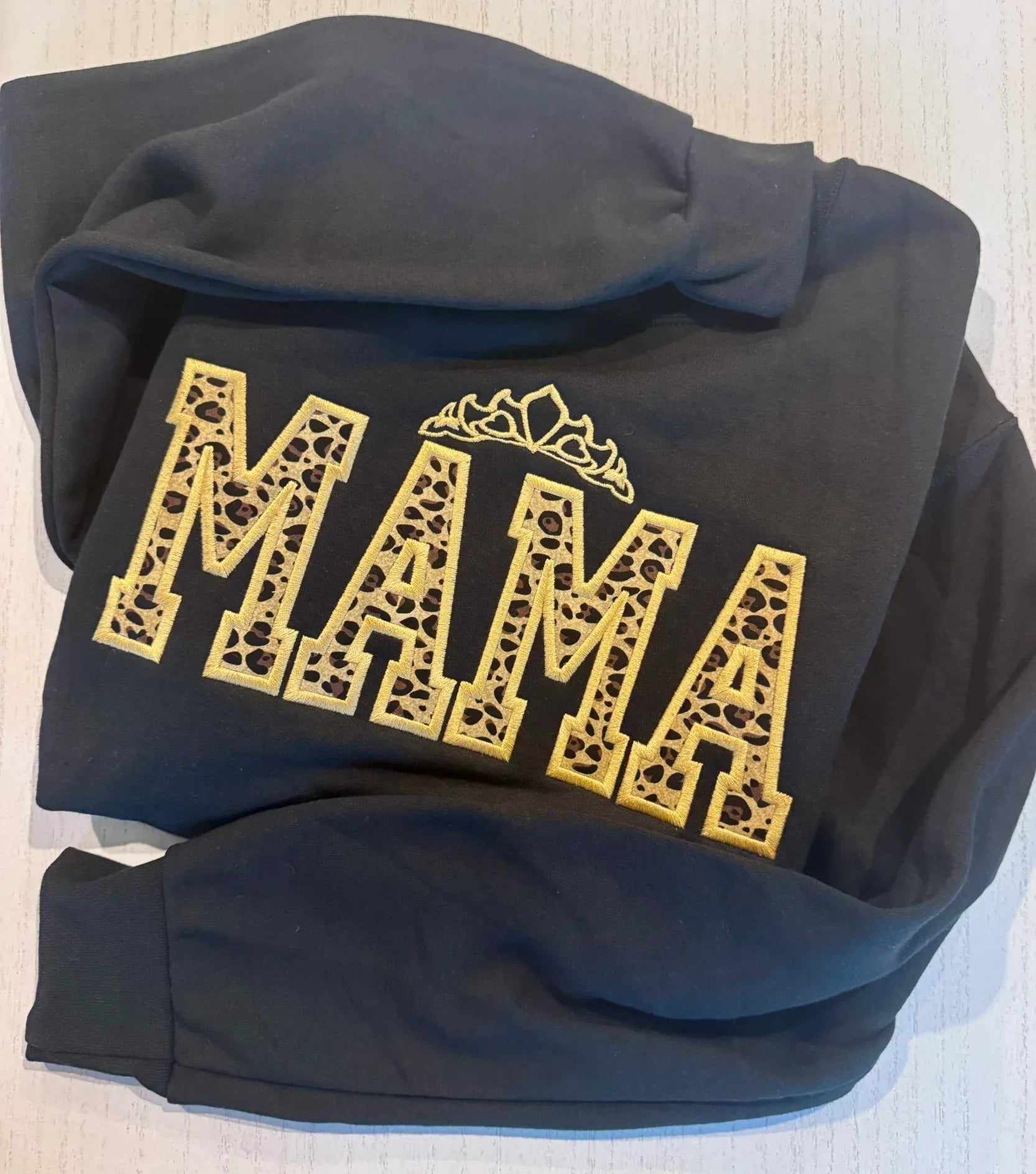 Black hoodie with 'MAMA' printed in leopard fabric and outline embroidered gold on a light background 
