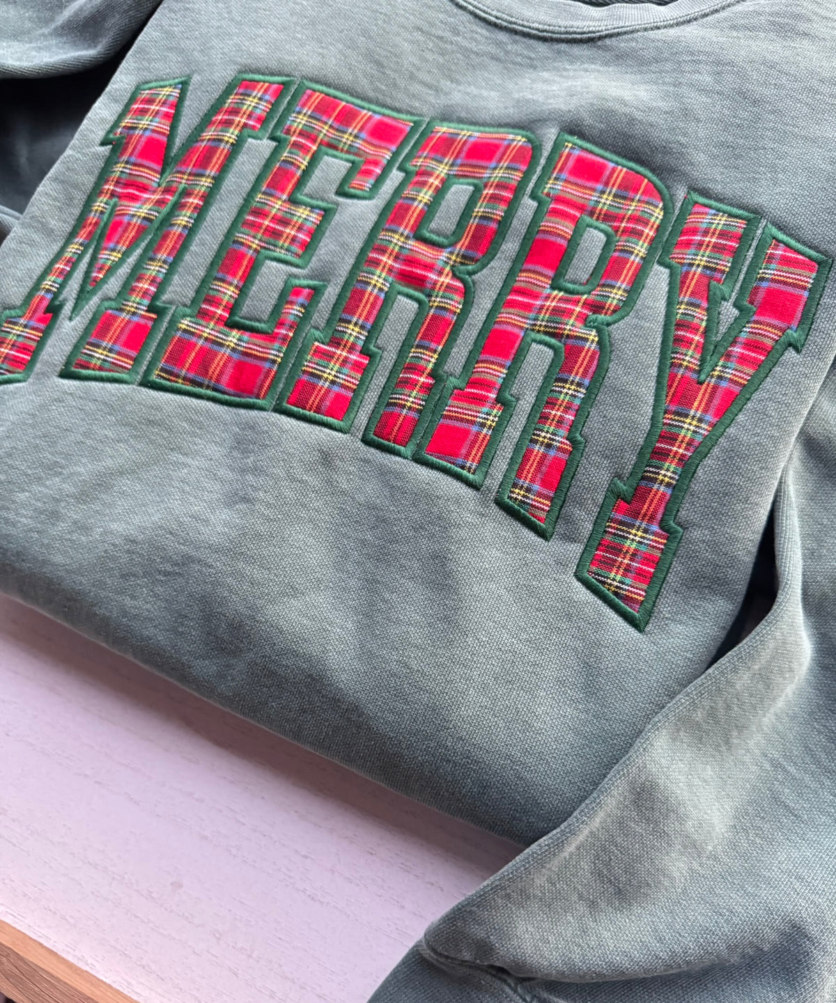 Close-up of the red plaid MERRY appliqué with green embroidered outline on a light green Christmas sweatshirt.