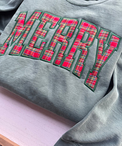 Close-up of the red plaid MERRY appliqué with green embroidered outline on a light green Christmas sweatshirt.