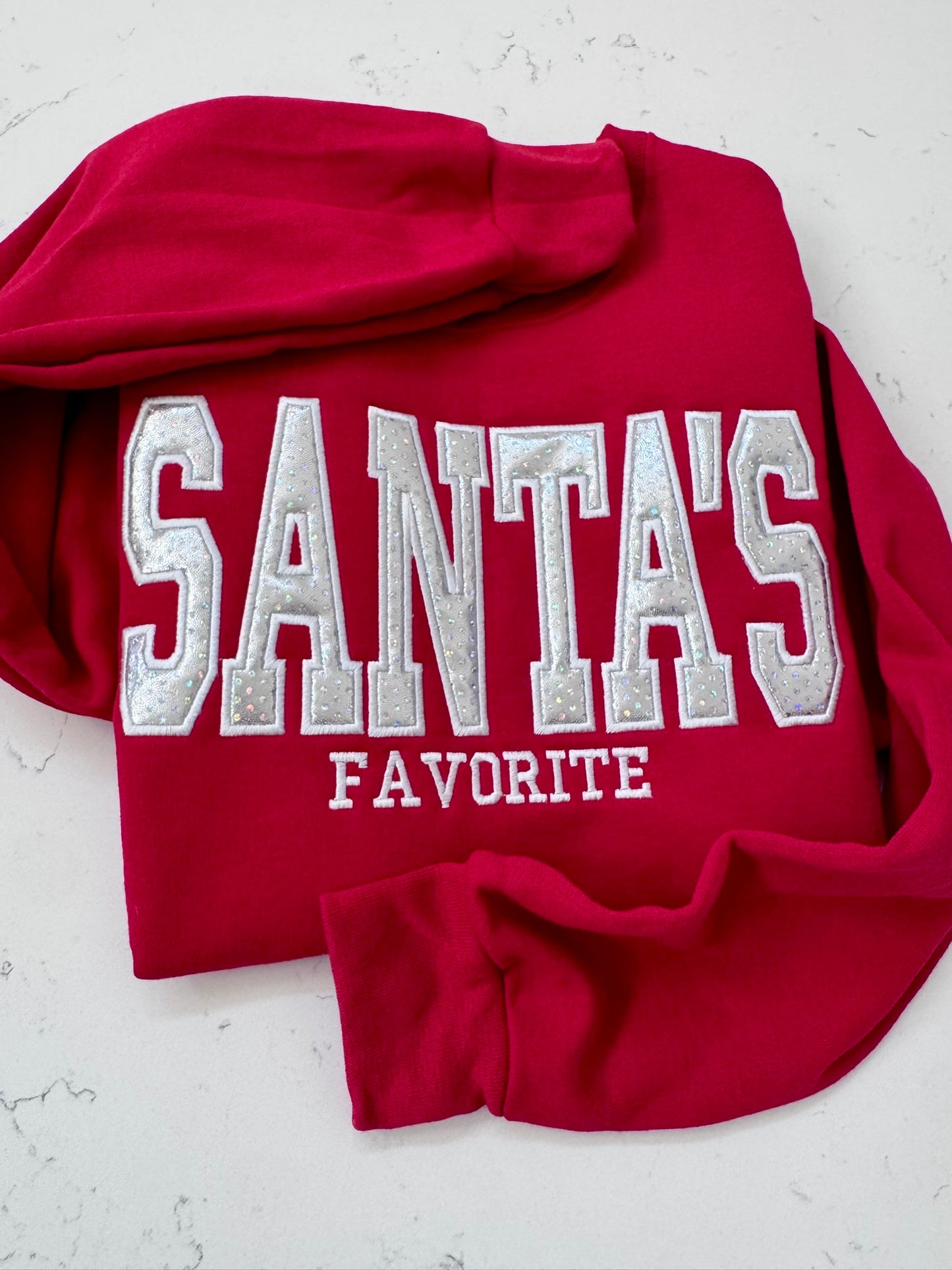A red hooded sweatshirt laid flat on a light textured surface, featuring large silver and white embroidered text that reads 'SANTA'S FAVORITE' across the front.