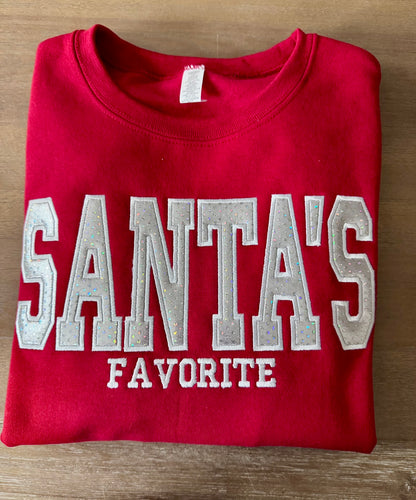 A folded red crewneck sweatshirt with 'SANTA'S FAVORITE' embroidered in large silver and white varsity-style letters on the front, laid flat on a wooden surface.