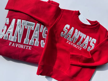 A red crewneck sweatshirt with 'SANTA'S FAVORITE' embroidered in distressed white block letters, shown folded and laid flat on a white background.