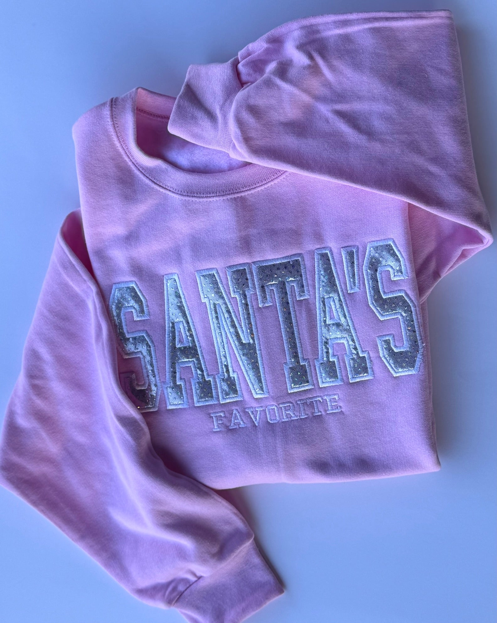 A folded light pink sweatshirt with 'SANTA'S FAVORITE' embroidered across the front in a glittery applique