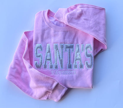 A folded light pink sweatshirt with 'SANTA'S FAVORITE' embroidered across the front in a glittery silver