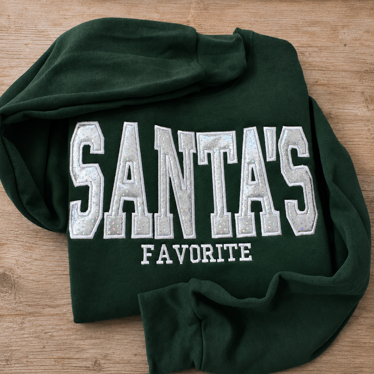A dark green fleece sweatshirt with 'SANTA'S FAVORITE' embroidered in white and silver on the front, laid flat on a light brown wooden surface.