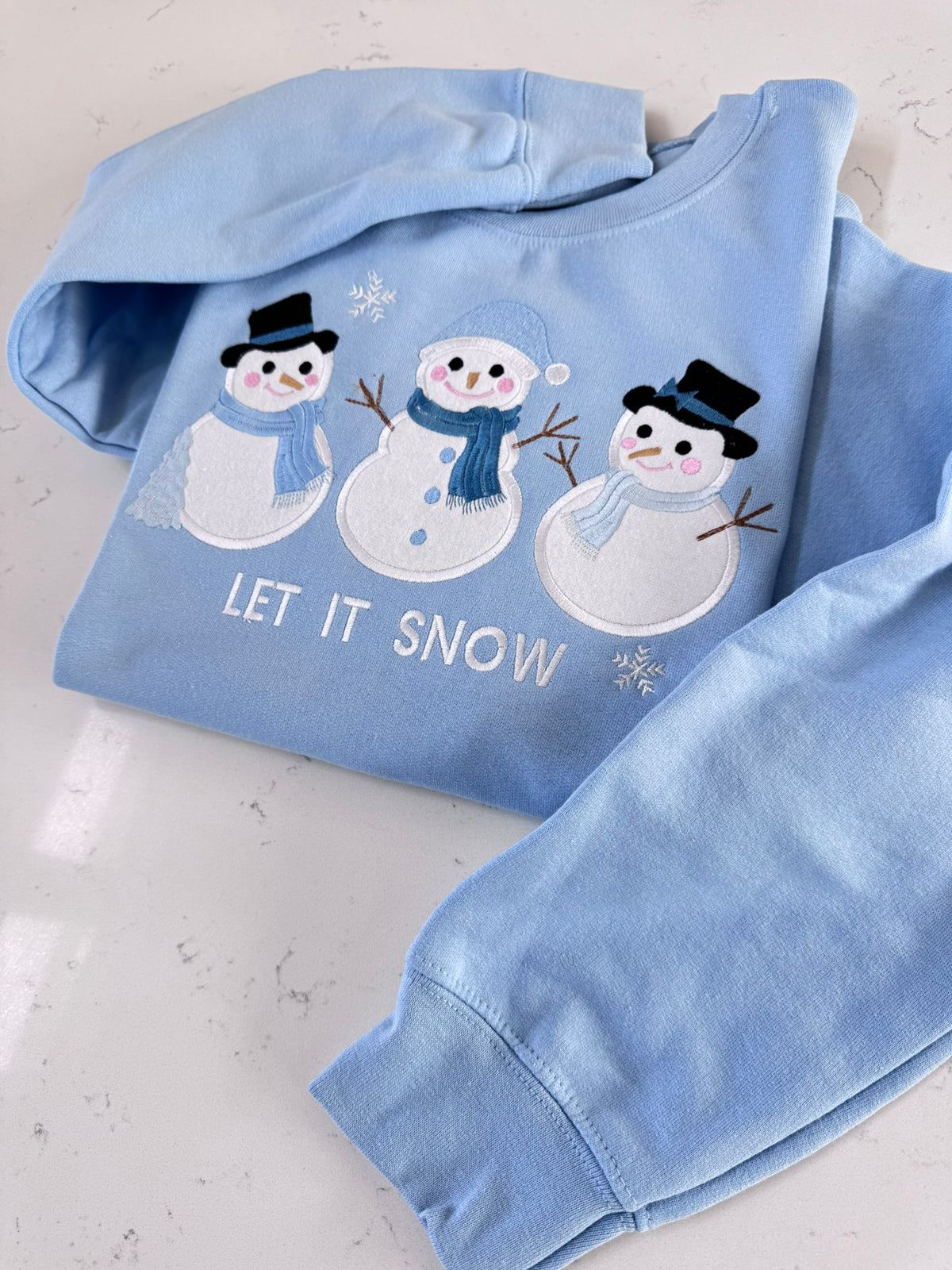 Folded light blue winter crewneck sweatshirt with three embroidered snowmen and Let It Snow lettering.