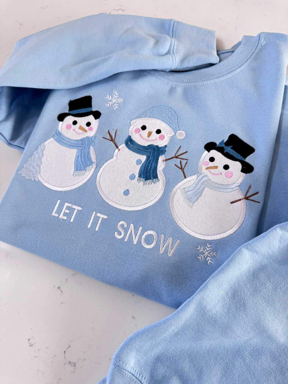 Detailed view of three snowmen with fabric appliqué, embroidered winter accessories and Let It Snow text.