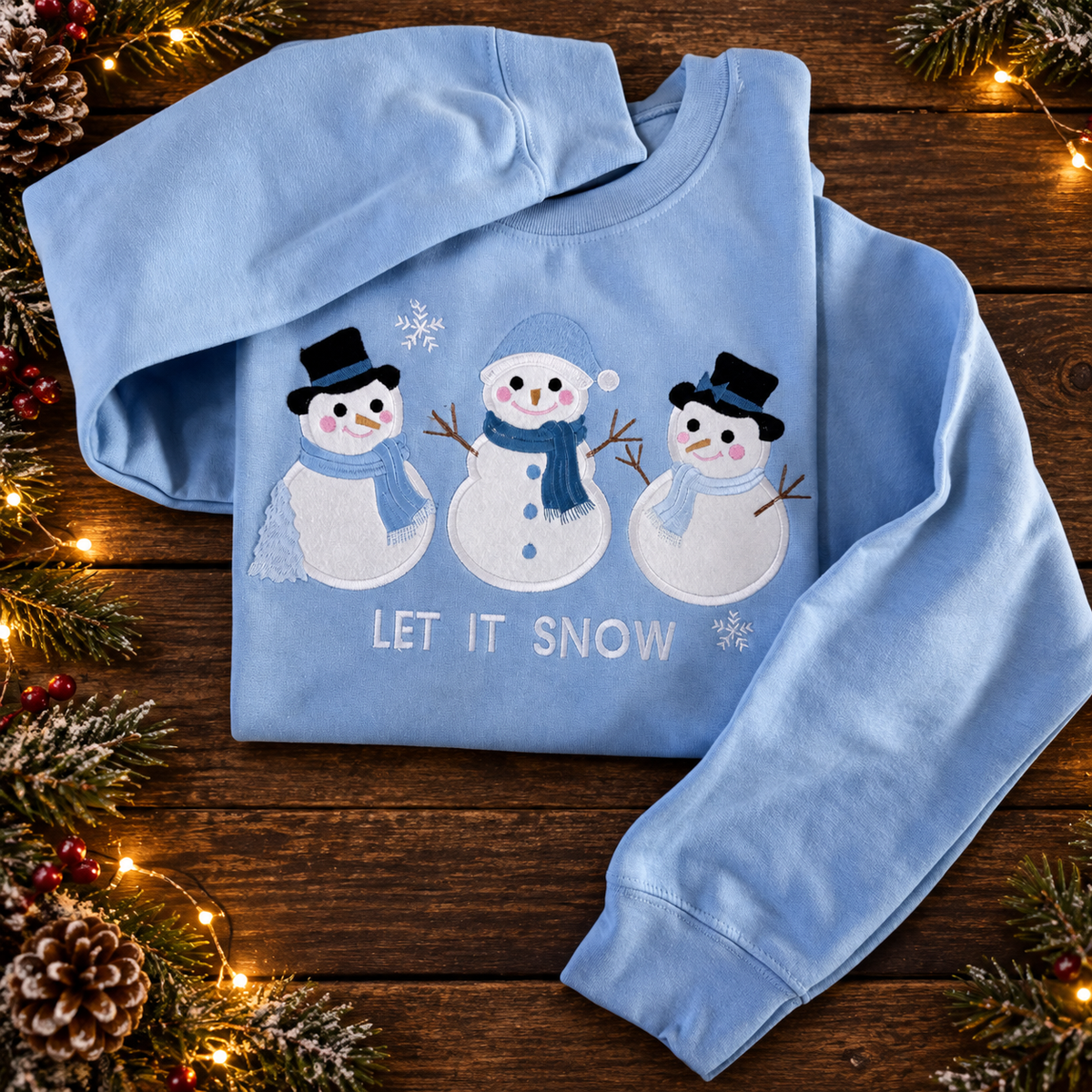 Blue embroidered snowman sweatshirt displayed on rustic dark wood with Christmas greenery and warm lights.