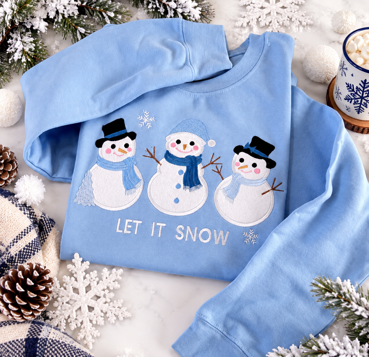 Light blue snowman sweatshirt surrounded by snowy pine branches, winter decorations and white snowflakes.