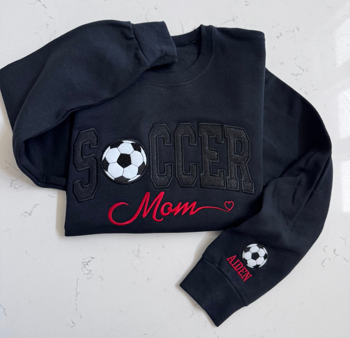 Folded black soccer sweatshirt featuring tonal sparkle and red Mom lettering
