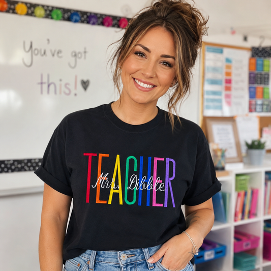 A smiling woman wearing a black graphic t-shirt with the word 'TEACHER' in rainbow colors and 'Mrs. Ebbie' in white script, standing in a classroom setting.