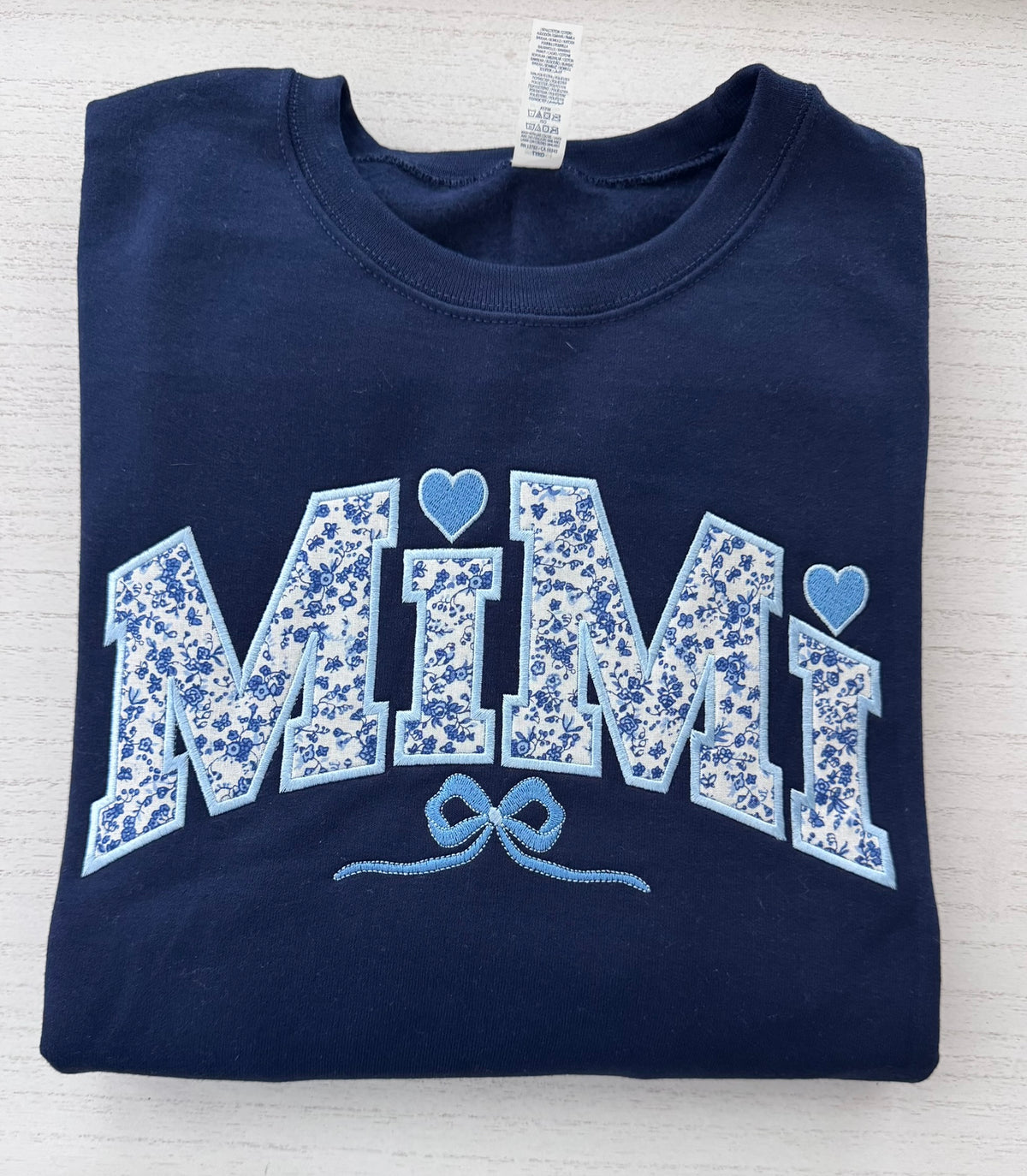 A folded navy blue t-shirt with 'MIMI' written in arched, light blue and white floral-patterned applique letters, accented with two light blue hearts and a bow design embroidered underneath, laid flat on a light wood background.