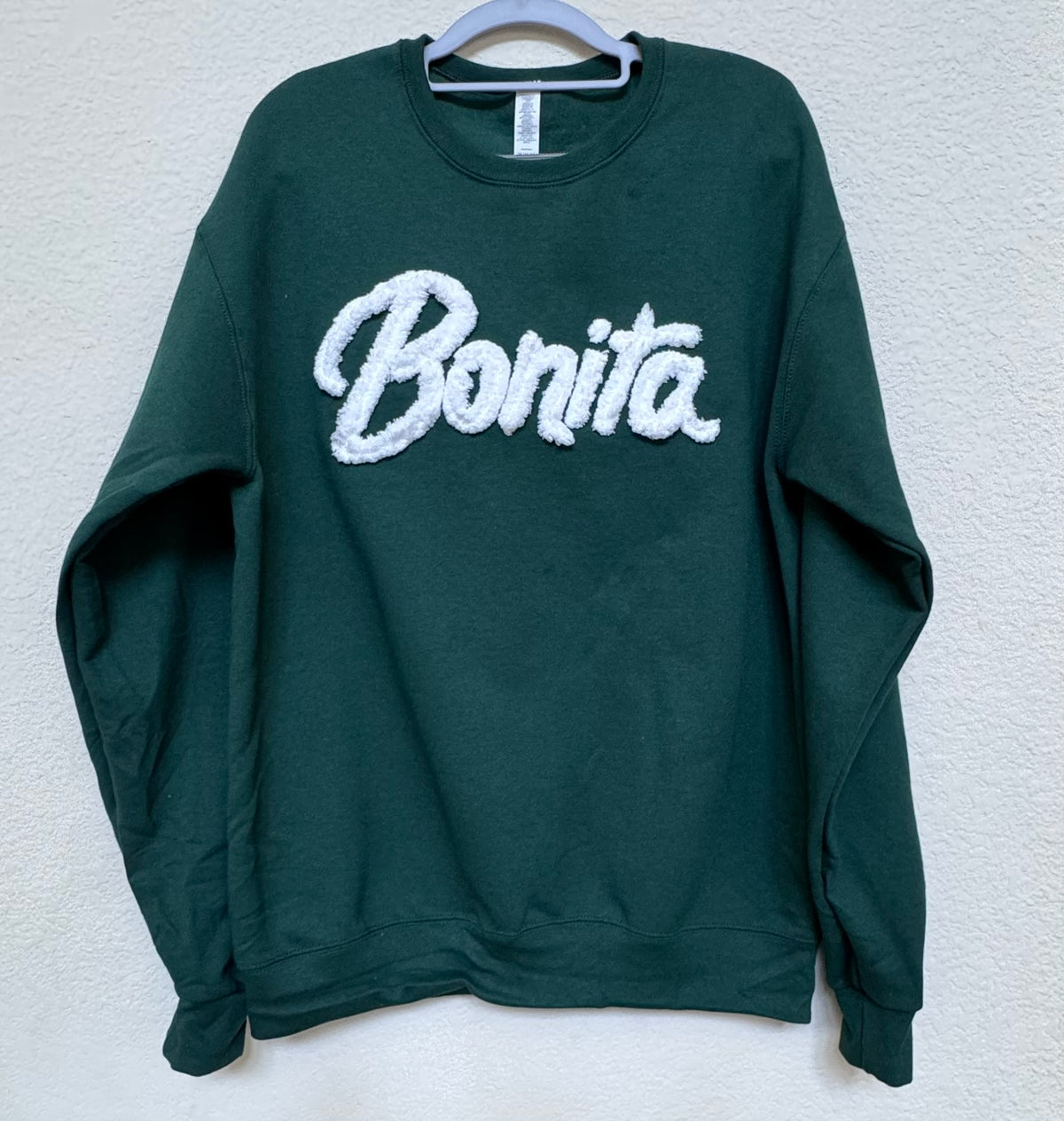 A dark green crewneck sweatshirt with 'Bonita' embroidered in white across the chest, hanging on a light blue hanger against a textured off-white wall.