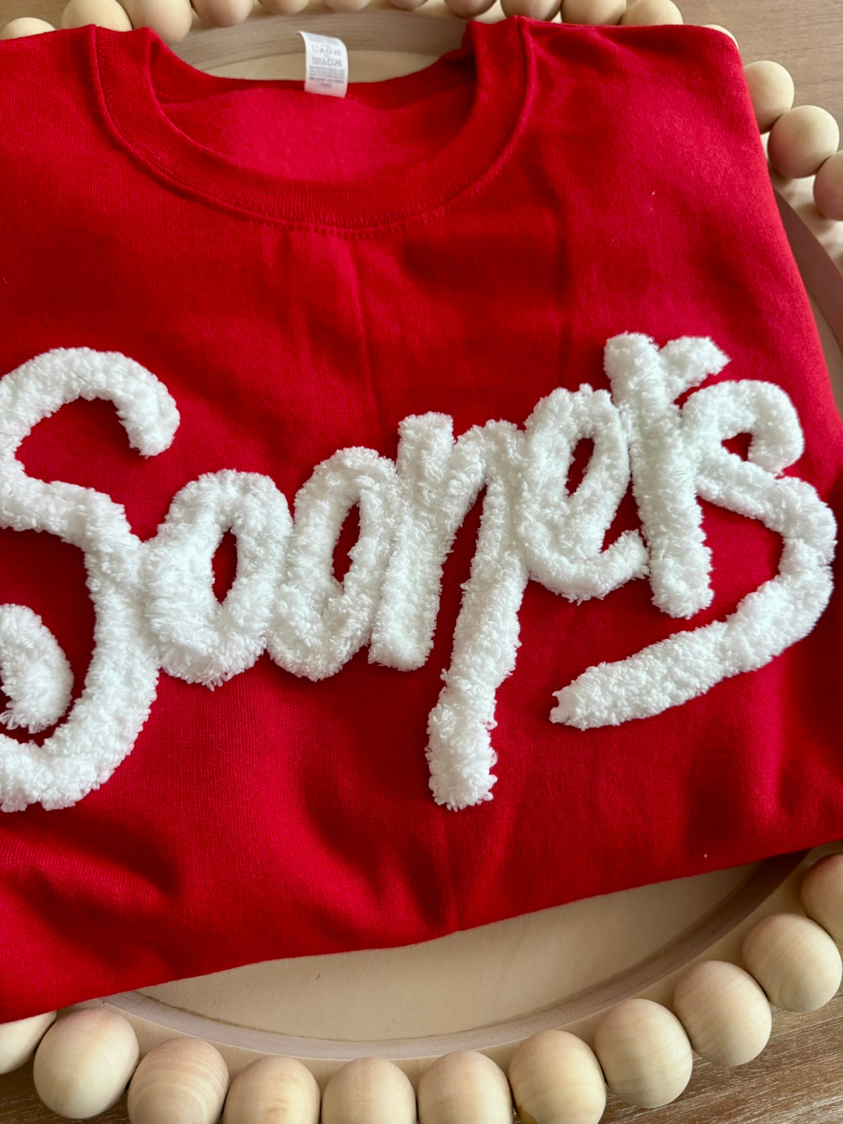 A folded red sweatshirt featuring a large, textured white fluffly-letter embroidery across the front, displayed on a wooden beads.
