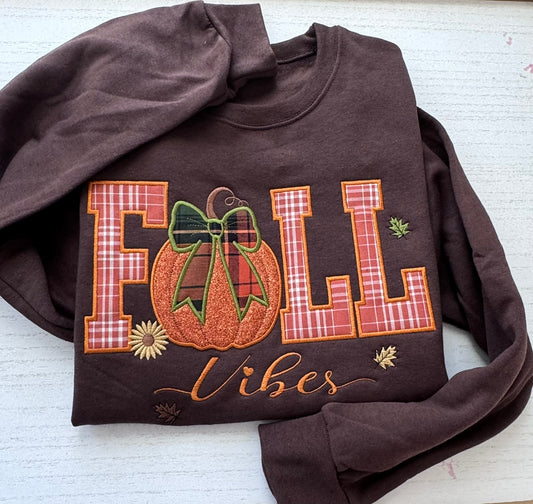 A dark brown crewneck sweatshirt laid flat on a white wood background, featuring a large 'FALL Vibes' embroidery design with a pumpkin and bow motif.